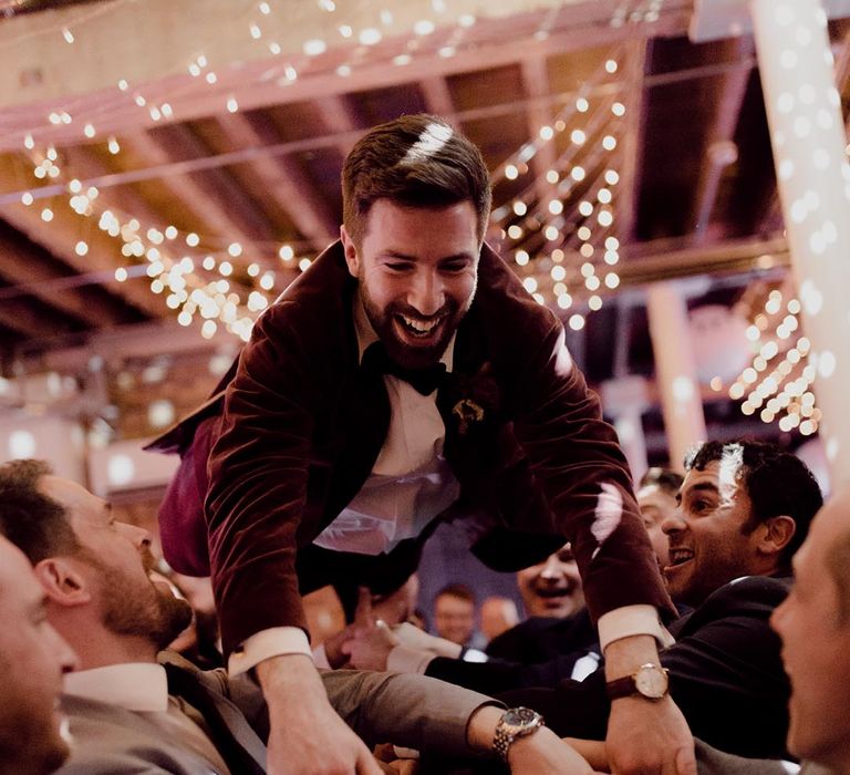 Groom wearing black bow tie and red suit jacket is lifted up in a dance by his groomsmen and other guests