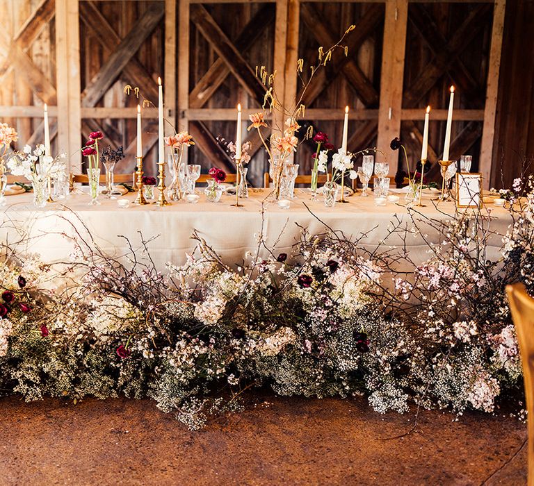 Large flower display along the top table at wedding reception with classic styling with white candles and tablecloth