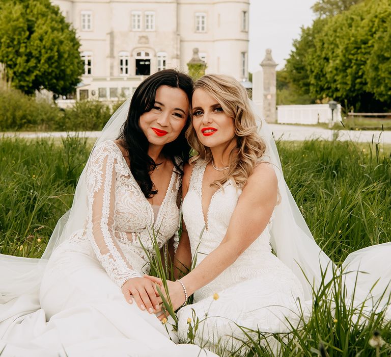 Brides in lace wedding dresses sit down together in the grass wearing matching bright red lipsticks 