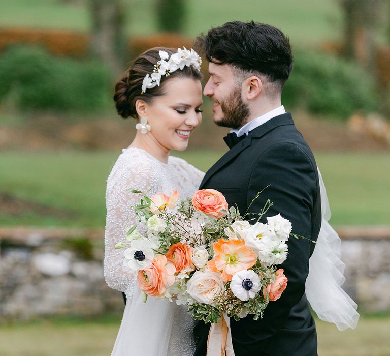 Groom in a tuxedo embracing his bride in a beaded wedding dres with smokey eyeshadow wearing a flower headdress and matching earrings as she holds a peach and white flower bouquet 