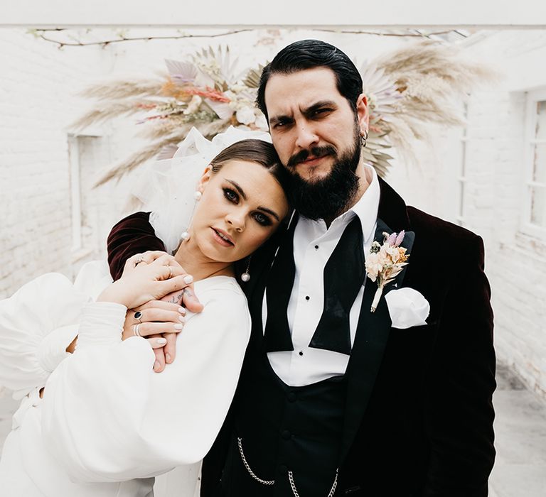 Beaded groom in a tuxedo with open bow tie embracing his bride in a long sleeve wedding dress and dangly earrings in a summer house 