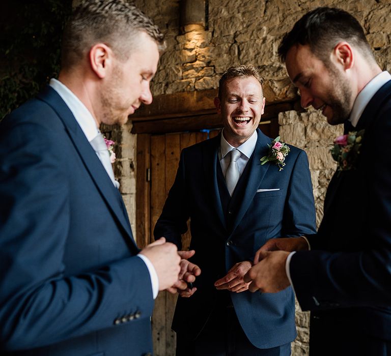 Groom laughs with groomsmen in blue suits and pale blue ties for wedding at Cripps Barn