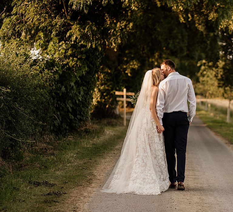 Bride and groom walk along a road holding hands and kissing 