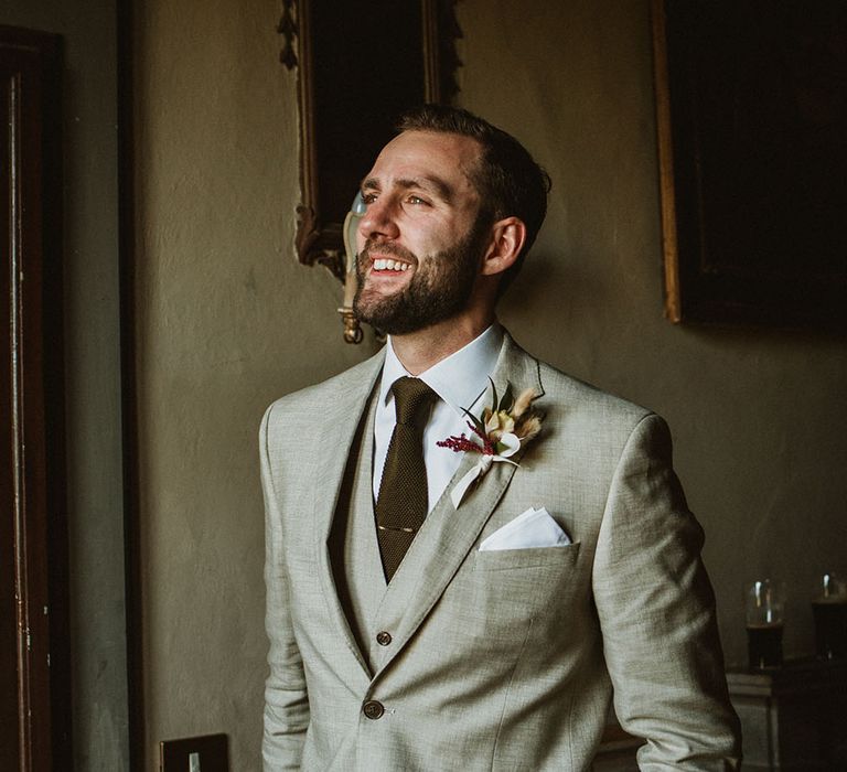 Groom wears three piece stone beige suit complete with green wool tie, gold tie pin and floral buttonhole 