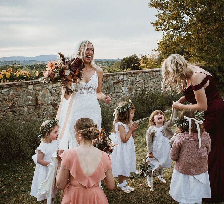 Bride laughs as she stands with her flower girls who wear white dresses and floral crowns whilst holding wicker baskets 