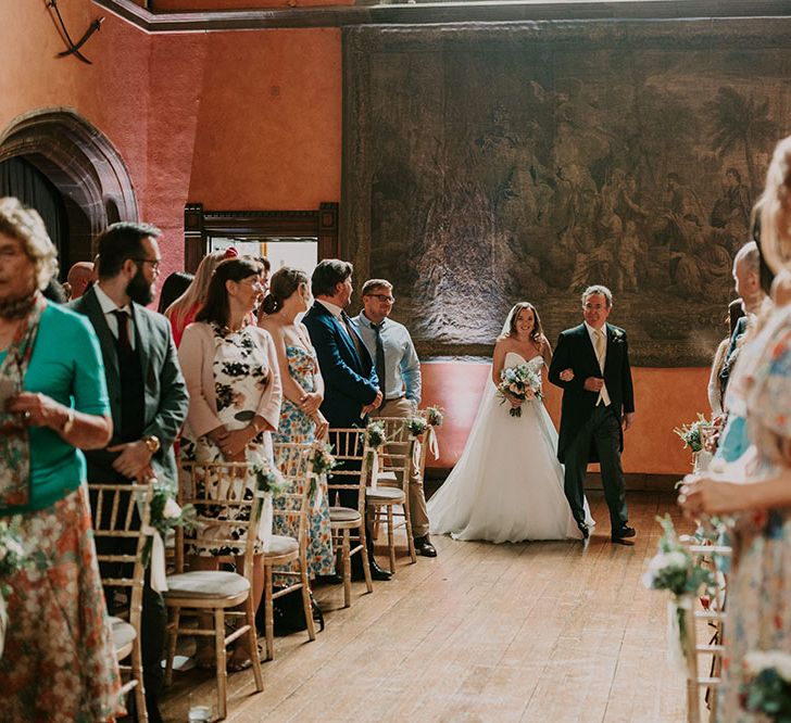 Bride walks down the aisle with her father at Cowdray House in front of tapestry wall hanging 