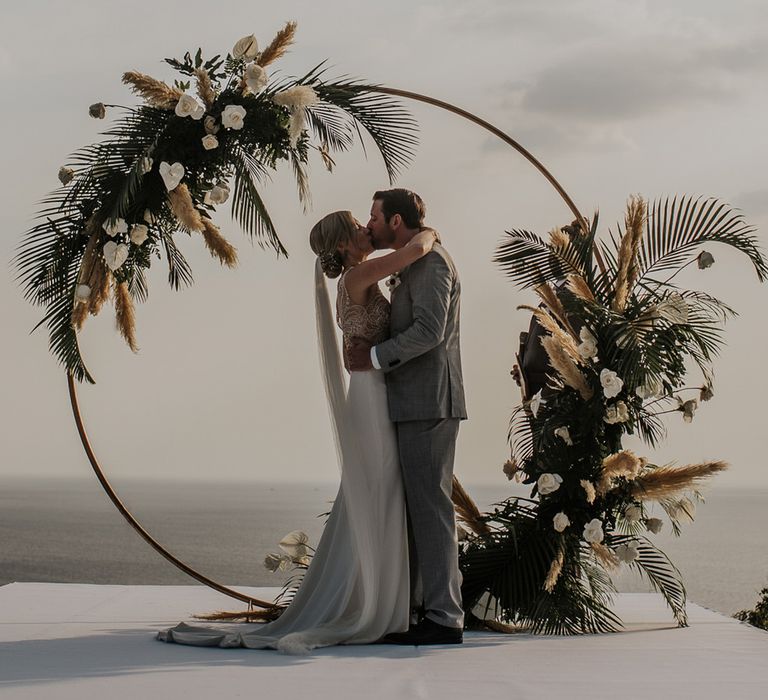 Bride and groom share a kiss in front of the moongate with fern leaves and pampas grass 