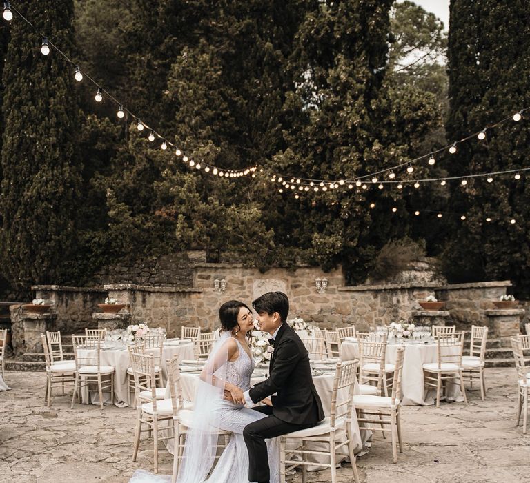 Bride & groom sit at table for chic and neutral outdoor wedding reception in Barcelona beneath strings of festoon lighting 