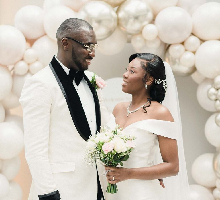 Bride wearing off-the-shoulder fitted wedding dress & groom in black tie stand in front of white and silver balloon arch