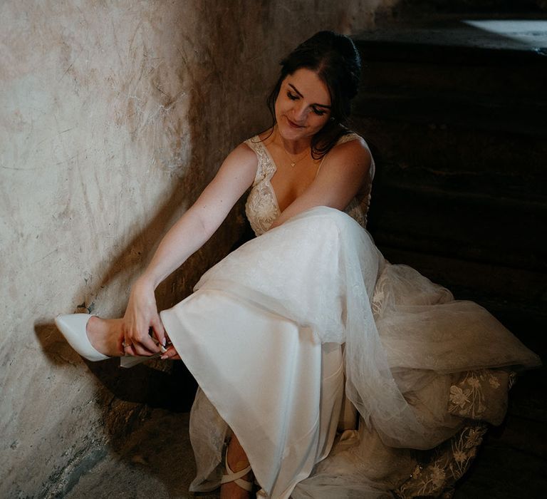 Bride wearing lace wedding dress removes her pointed satin shoes on her wedding day