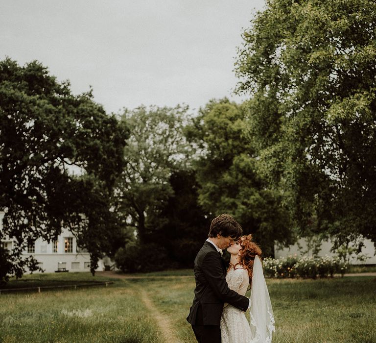 Bride wears her red hair in curls complete with lace edged veil as she stands with her groom in front of Belair House 