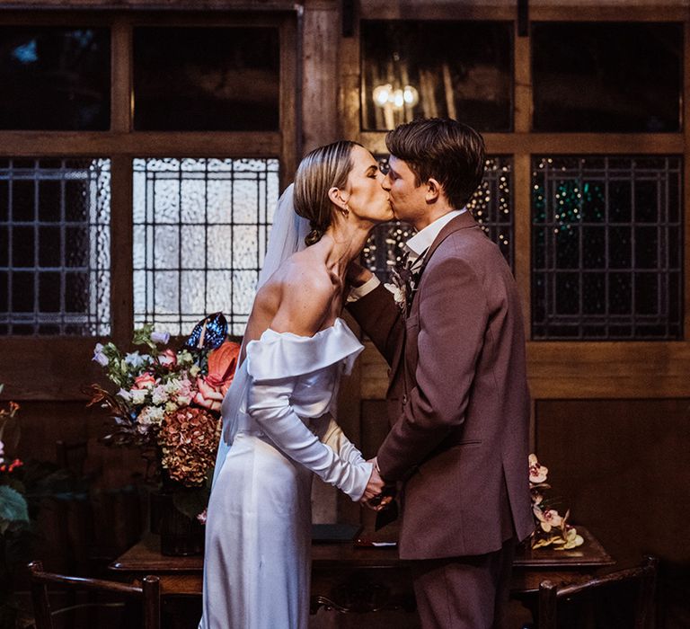 Bride in an off the shoulder satin cowl back wedding dress shares the first kiss with the groom in a brown suit 