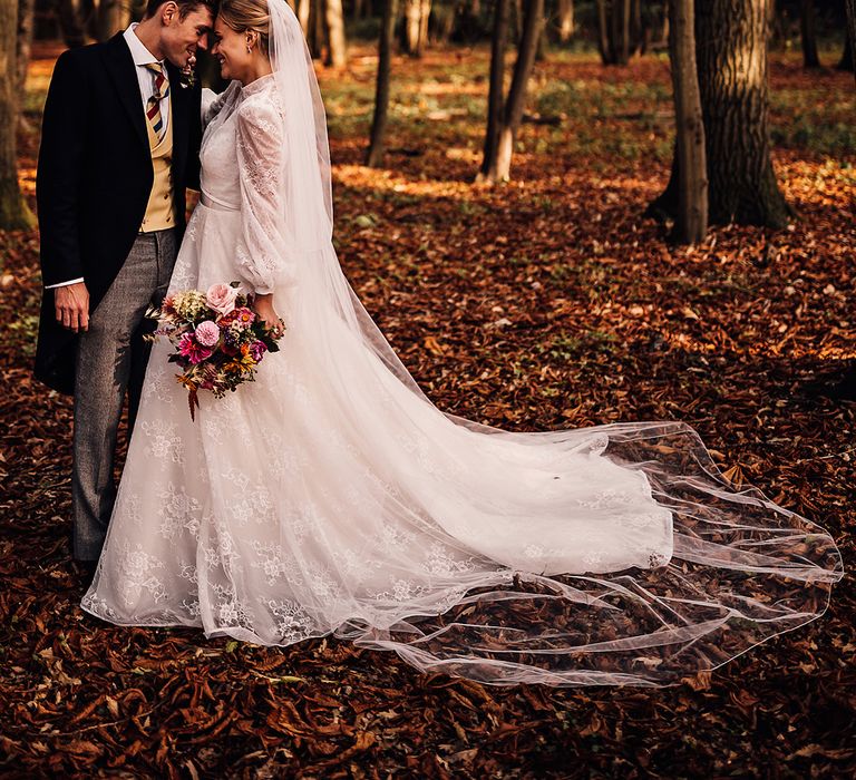 Groom in a morning suit puts his forehead against the bride who wears a lace wedding dress in the woods 