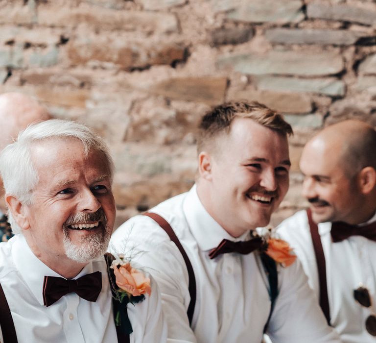 Groomsmen sit in their white shirts and burgundy braces and bow ties for the ceremony 