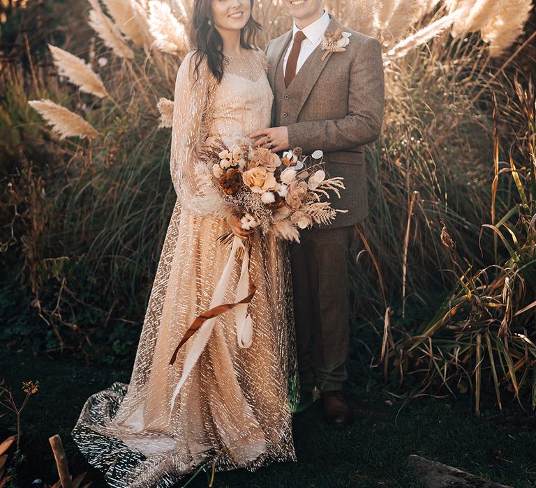 Bride in gold wedding dress and her groom stand in front of large pampas grass outdoors