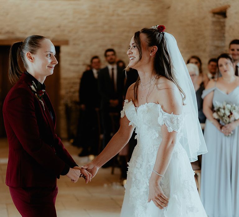 Bride in an off the shoulder wedding dress standing at the altar with the bride in a burgundy suit 