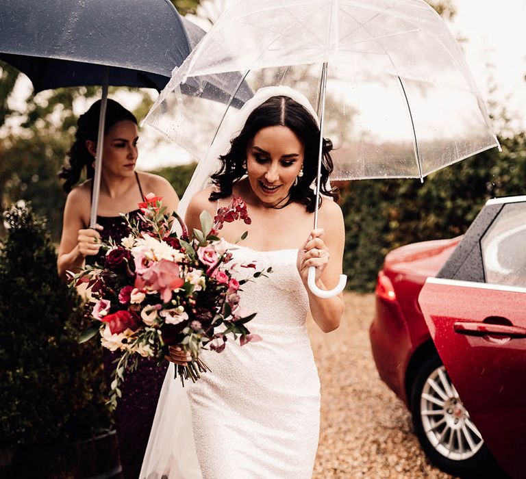 The bride holds a clear umbrella as she walks in a beaded sparkle wedding dress and pearl wedding shoes to the ceremony 