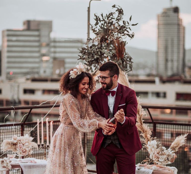 Bride in sparkly wedding dress and groom in red suit doing a champagne pop 