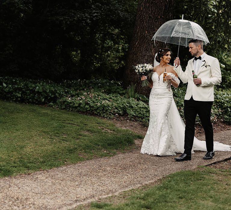 Bride in a floral lace off the shoulder wedding dress walks with the groom in a white suit jacket under an umbrella 