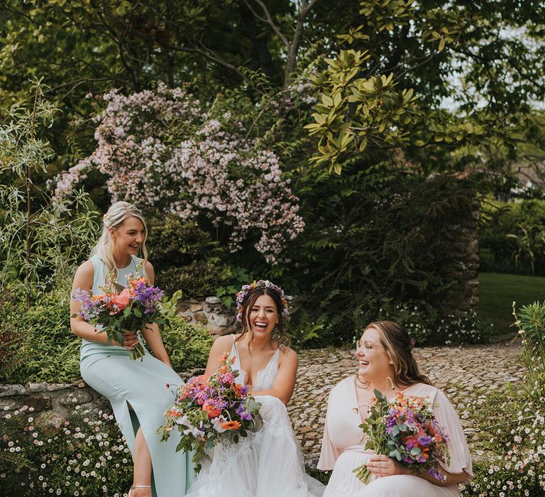 Bride sits with her bridesmaids in mismatched dresses holding colourful floral bouquets 