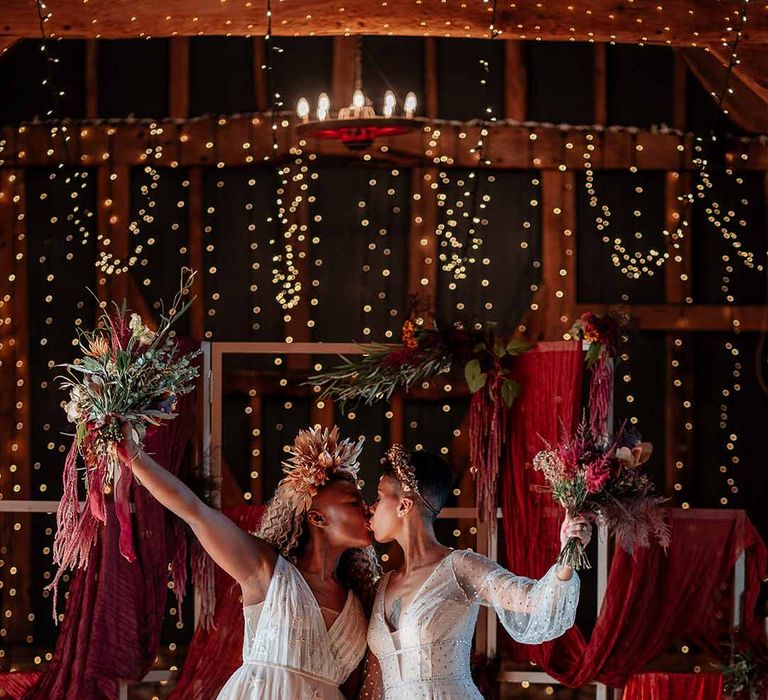 Bride in sleeveless floaty peach gown and gold leaf crown kissing bride in crystal embellished gown and gold headdress with red wedding drapes, foliage and dried flower decor and fairy lights in the background