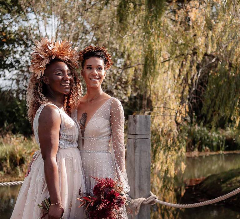 Bride in sleeveless floaty peach gown and gold leaf crown and crystal embellished gown and gold headdress holding autumnal dried flower bouquets standing by the river at Silchester farm 