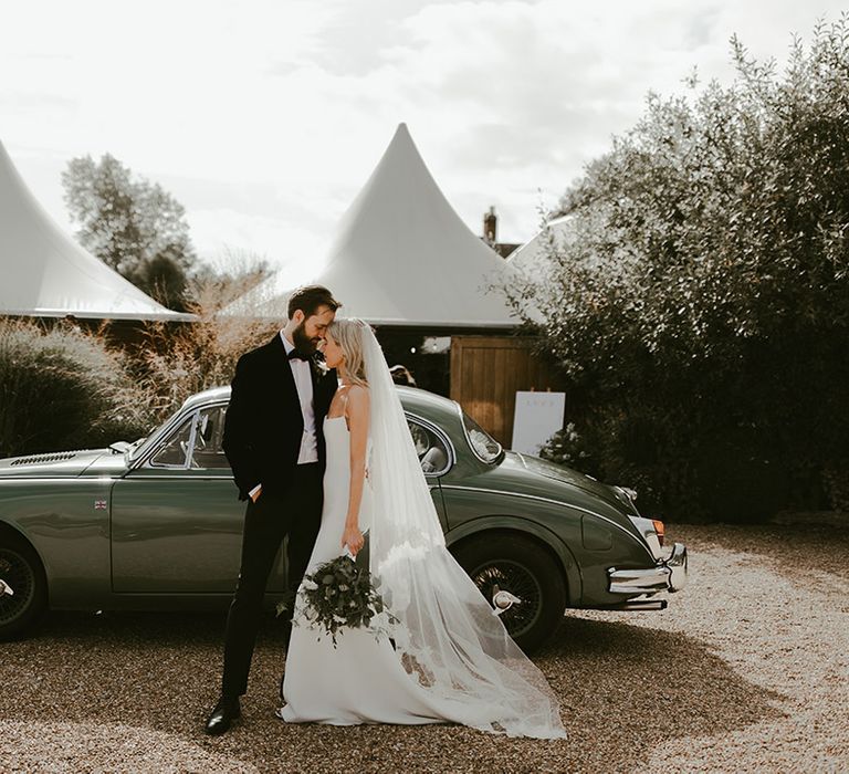 The bride and groom rest their foreheads against each other as they stand next to their green vintage wedding car