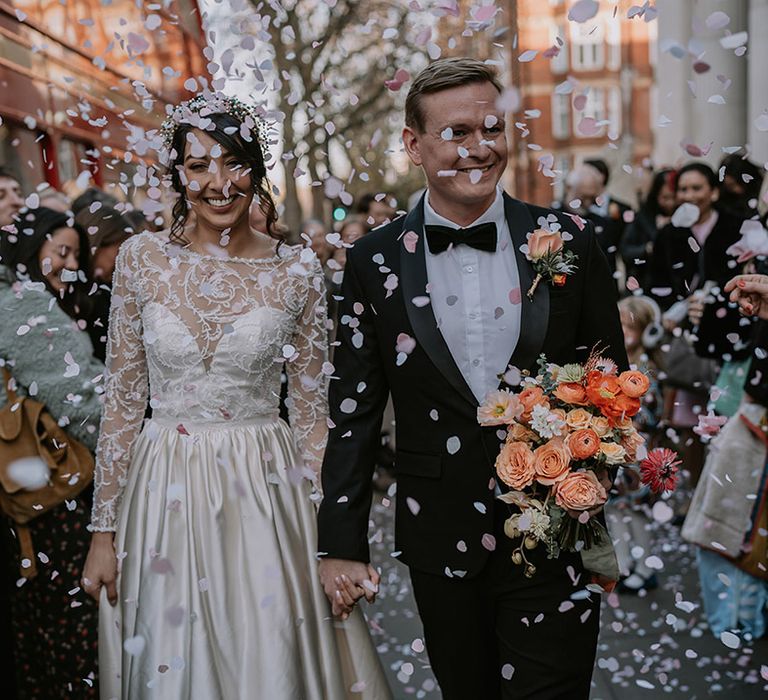 Bride in long sleeve lace wedding dress and groom in classic black tux doing confetti walk at Christmas multicultural London wedding