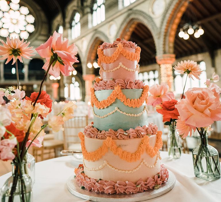 Brightly coloured retro styled three tier wedding cake complete with ruffled icing beside colourful floral arrangements 