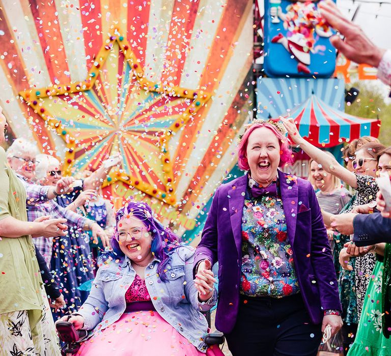 Bride in purple velvet blazer and black suit trousers with purple bowtie, pink patterned pocket square and colourful patterned shirt holding hands with bride in wheelchair wearing personalised denim jacket, pink wedding dress and blue tights doing colourful confetti throw 