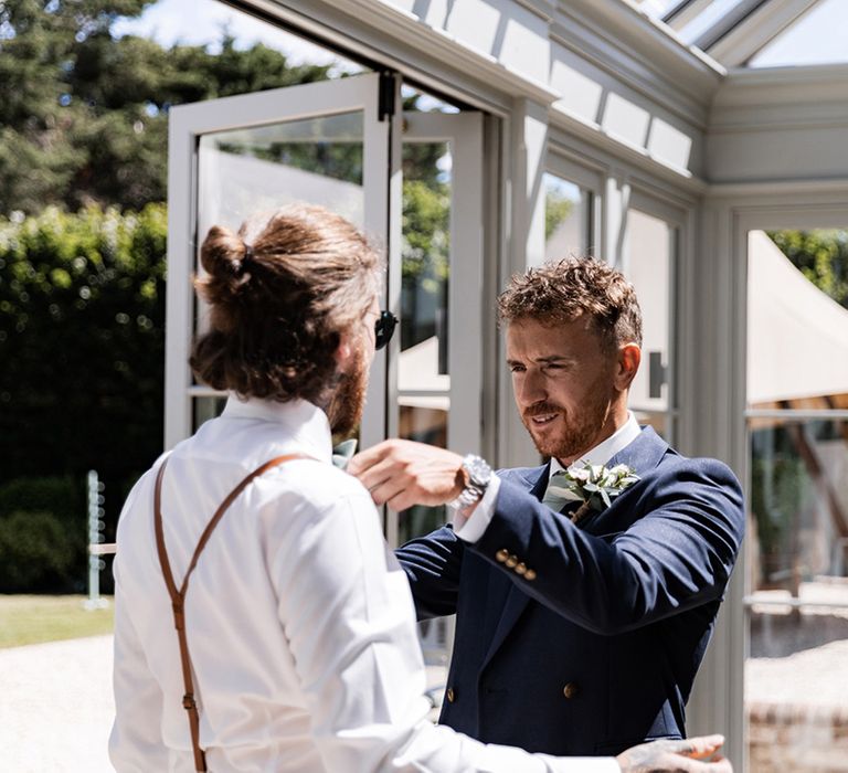 Groom in double breasted blazer helps groomsmen get ready on the morning of his wedding day