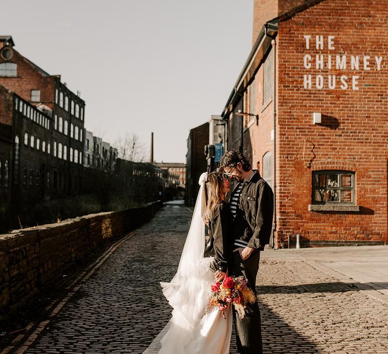 Bride in ruched church length wedding veil, wedding dress and personalised black leather jacket holding colourful bridal bouquet and groom in striped long sleeve t shirt and black denim jacket standing outside Sheffield wedding venue 