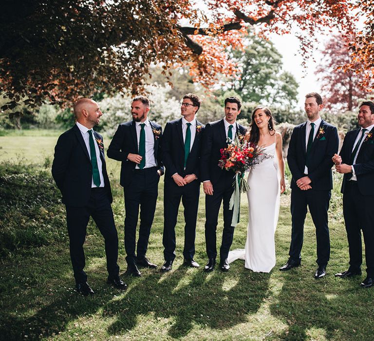 Bride and groom stand laughing with the groomsmen in matching navy suits and green ties for the wedding 