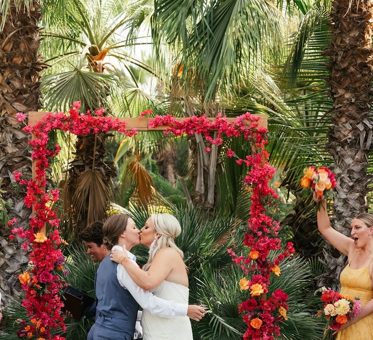 Brides kiss beneath red floral archway outdoors during ceremony in Mexico 