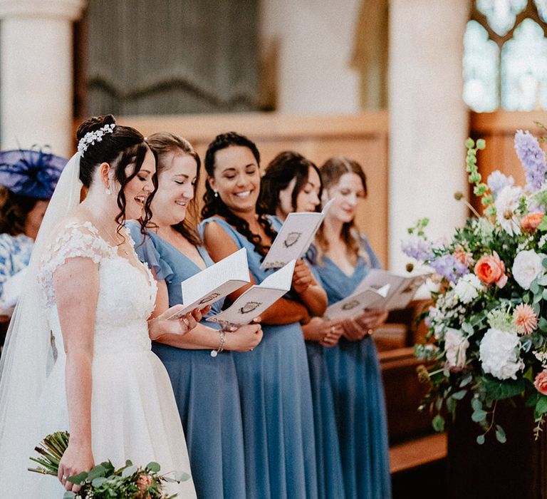 The bride stands with her bridesmaids in blue bridesmaid dresses reading out the hymns for the church wedding ceremony 