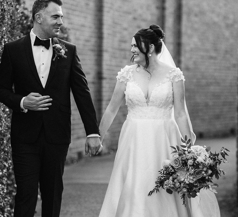 The bride wears a flower wedding dress with her brown hair in an elegant updo walking with the groom in a black tuxedo 