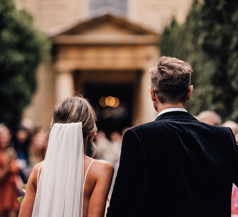 Groom in black velvet tuxedo holding hands with the bride in a fitted lace Grace Loves Lace wedding dress 