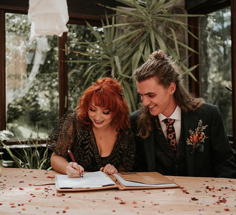 Groom in dark grey three piece suit and burgundy flower patterned tie and bride in black dress for wedding signing  