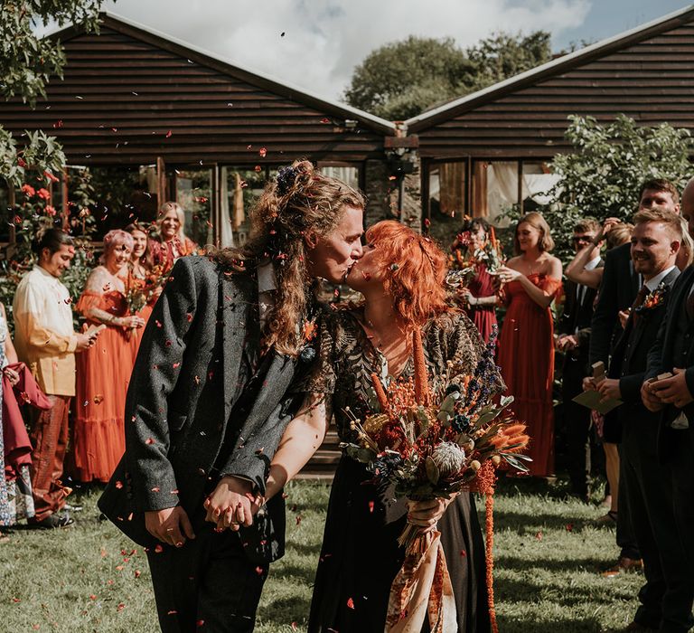 Groom in dark grey three piece suit and burgundy flower patterned tie and bride in embellished black wedding dress holding large orange dried flower bouquet kissing during wedding confetti exit 