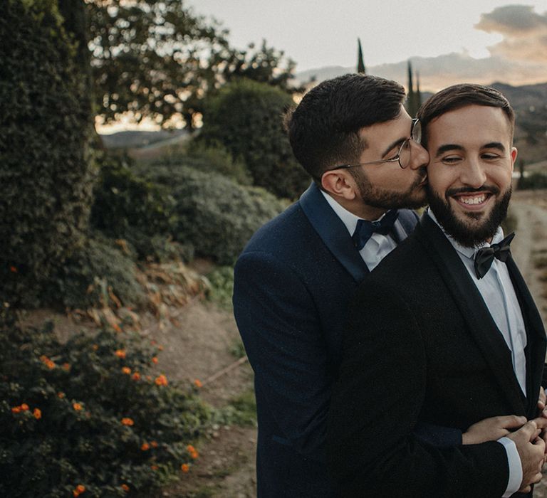 Groom in a navy blue suit kissing his groom in a black tuxedo and bow tie 