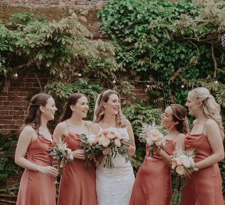 The bride laughs with her bridal party who wear matching satin cowl neck midi coral bridesmaid dresses for the rustic luxe wedding 