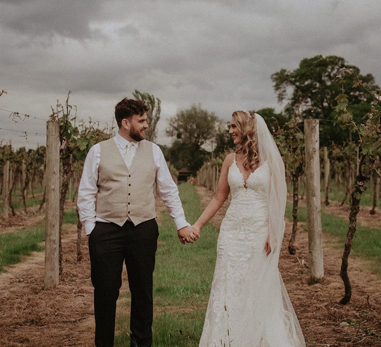 Groom in black trousers wearing a white shirt with beige waistcoat stand outside in the grounds of Stanlake Park with the bride in her fitted lace wedding dress 