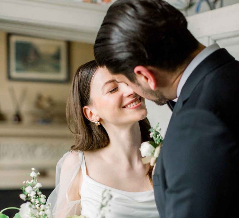 Groom in classic black tuxedo and boutonniere embracing bride in long sheer puff sleeve satin wedding dress with cowl neck