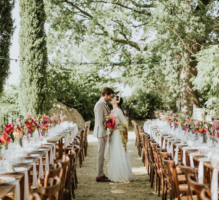 Bride in lace wedding dress and bridal crown with a pink bridal bouquet kissed groom in greige suit surrounded by wedding tables covered in pink floral decor