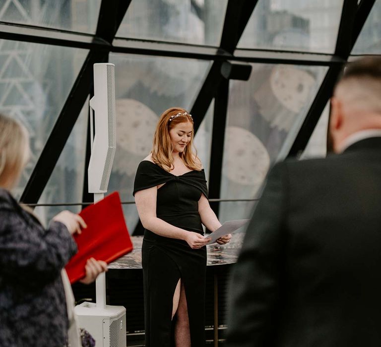 Wedding guest wearing velvet black off the shoulder bridesmaid dress with front leg slit doing wedding speech at The Gherkin wedding venue London