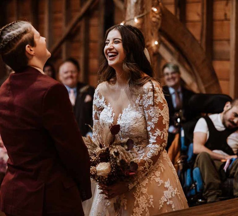 Bride in lace illusion sleeve wedding dress with overlay holding large autumnal boho dried flower bouquet laughing at the alter with bride in maroon bridal suit 