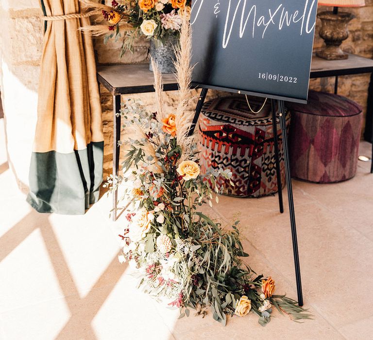 Black wedding welcome sign on wooden easel with autumnal flower arrangement with orange flowers and pampas grass 