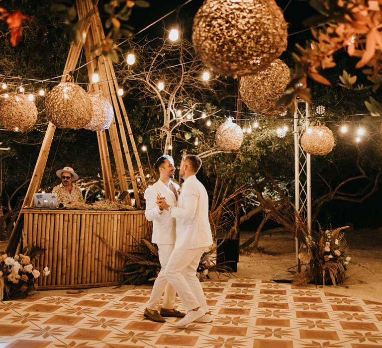 Grooms in matching white grooms suits doing their first dance as a couple on DIY hand painted wedding dance floor under twine lampshades and fairy lights 