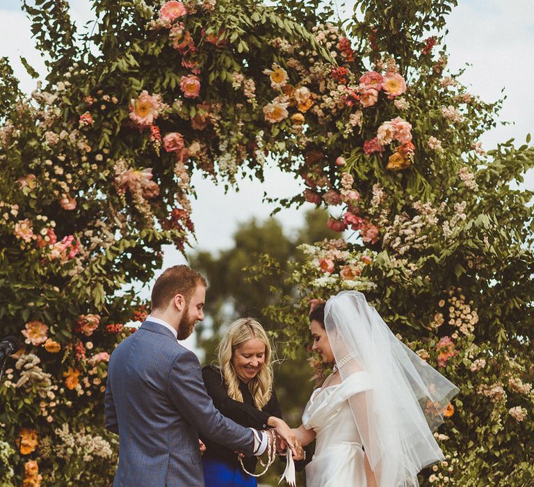 Wedding celebrant performs bespoke handfasting ceremony for the bride and groom at their outdoor wedding with incredible floral arch decoration 