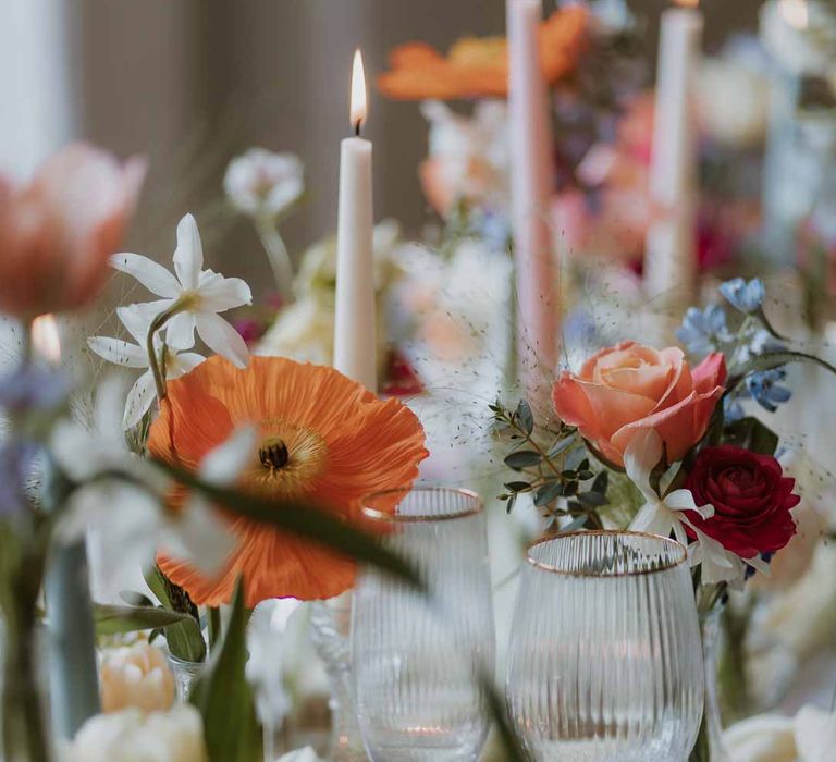 Spring themed wedding tablescape with orange poppies, pink and red roses, jasmine and bluebells on white wedding tablecloth and off-white satin table runner with pastel tapered candles, gold-rimmed wine glasses and classic crockery 